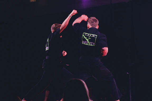 Two individuals are performing martial arts on a dimly lit stage. They are wearing black uniforms with neon green logos on the back labeled 'Team Infinity'. One person is throwing a punch while the other is sidestepping or responding to the movement. The focus is on dynamic action and physical combat training.