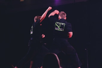 Two individuals are performing martial arts on a dimly lit stage. They are wearing black uniforms with neon green logos on the back labeled 'Team Infinity'. One person is throwing a punch while the other is sidestepping or responding to the movement. The focus is on dynamic action and physical combat training.