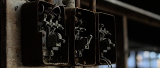 A close-up view of three old, rusty electrical fuse boxes mounted on a wooden board. The metal casings show signs of deterioration, and wires are visible inside each box. The environment appears to be dimly lit, emphasizing the aged condition of the fuse boxes.