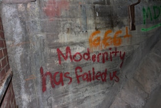 Graffiti is displayed on a rough concrete wall, featuring the phrases '666+' in orange and 'Modernity has failed us' in red. The setting appears aged and worn, with some sections of brickwork visible.