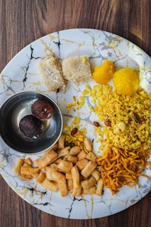 The image features a marble-patterned plate with various Indian snacks neatly arranged. There are two pieces of coconut burfi, two yellow-colored round sweets, a portion of flattened rice flakes mixed with nuts and spices, a pile of small fried dough sticks, and a serving of sev. A small metal bowl containing two dark brown gulab jamuns is also placed on the plate.