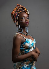 A person wearing a colorful headscarf with intricate patterns. They have embellishments on their face and are dressed in a floral-patterned dress. The lighting highlights their features against a plain background.