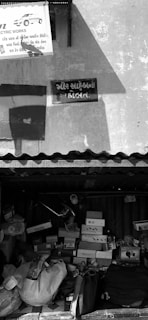 A small, cluttered shop front with various boxes and items piled up underneath a corrugated metal roof. The walls feature two signs, one of which is partially visible, advertising electrical works, while the other is in a different script. Shadows of nearby structures are cast on the wall.