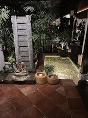 A serene setting featuring a golden Buddha statue placed on a stone pedestal surrounded by lush green plants. There are two candles in yellow holders on the tiled floor next to the statue. In the background, there's a small pond with clear water and pebbles, enhanced by soft ambient lighting. The environment conveys tranquility and calmness.