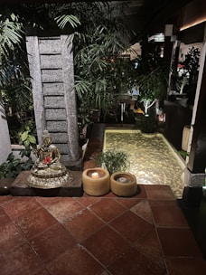 A serene setting featuring a golden Buddha statue placed on a stone pedestal surrounded by lush green plants. There are two candles in yellow holders on the tiled floor next to the statue. In the background, there's a small pond with clear water and pebbles, enhanced by soft ambient lighting. The environment conveys tranquility and calmness.