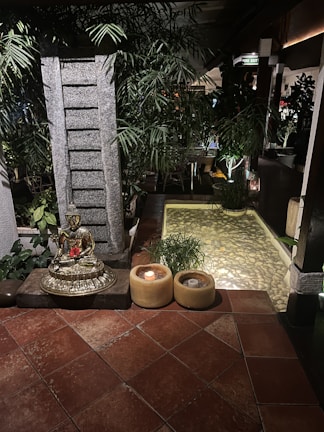 A serene setting featuring a golden Buddha statue placed on a stone pedestal surrounded by lush green plants. There are two candles in yellow holders on the tiled floor next to the statue. In the background, there's a small pond with clear water and pebbles, enhanced by soft ambient lighting. The environment conveys tranquility and calmness.