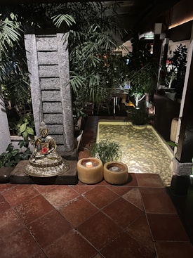 A serene setting featuring a golden Buddha statue placed on a stone pedestal surrounded by lush green plants. There are two candles in yellow holders on the tiled floor next to the statue. In the background, there's a small pond with clear water and pebbles, enhanced by soft ambient lighting. The environment conveys tranquility and calmness.