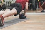 A person is performing a plank exercise on a mat, using a medicine ball for added balance and difficulty. The individual is dressed in athletic wear, including a red shirt and black shorts. This activity is taking place in a gym setting, as indicated by the mirrored wall and various exercise equipment in the background.