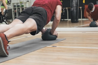 A person is performing a plank exercise on a mat, using a medicine ball for added balance and difficulty. The individual is dressed in athletic wear, including a red shirt and black shorts. This activity is taking place in a gym setting, as indicated by the mirrored wall and various exercise equipment in the background.