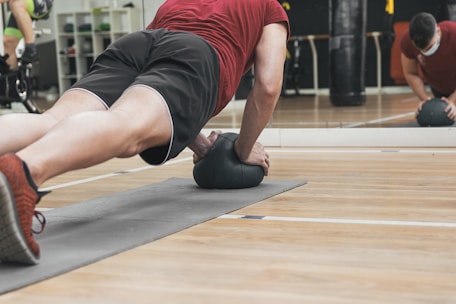A person is performing a plank exercise on a mat, using a medicine ball for added balance and difficulty. The individual is dressed in athletic wear, including a red shirt and black shorts. This activity is taking place in a gym setting, as indicated by the mirrored wall and various exercise equipment in the background.