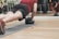 A person is performing a plank exercise on a mat, using a medicine ball for added balance and difficulty. The individual is dressed in athletic wear, including a red shirt and black shorts. This activity is taking place in a gym setting, as indicated by the mirrored wall and various exercise equipment in the background.