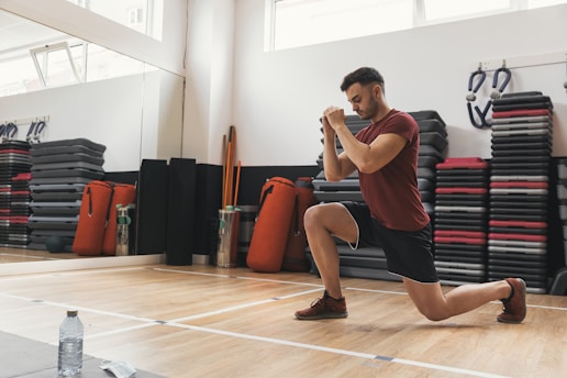 A man is performing a lunge exercise in a gym setting. He is wearing a red shirt and black shorts, focusing intently on holding his position. The background shows stacked exercise mats, wall-mounted exercise equipment, and several red punching bags. A large mirror reflects the gym space, and a water bottle is placed on the floor.