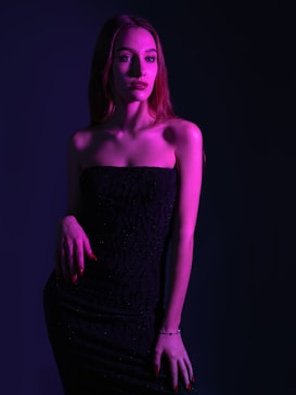 A woman stands confidently wearing a strapless black dress. The lighting casts a vibrant purple and pink hue across her, creating dramatic shadows. Her long hair cascades down her shoulders, and she has a poised, calm expression.