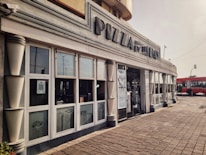 A restaurant with a streamlined facade featuring large windows and an art deco style. The signage reads 'PIZZA by the BAY,' and there is a take-away counter visible inside. The setting includes a tiled walkway and a red bus visible in the background, on a slightly overcast day.