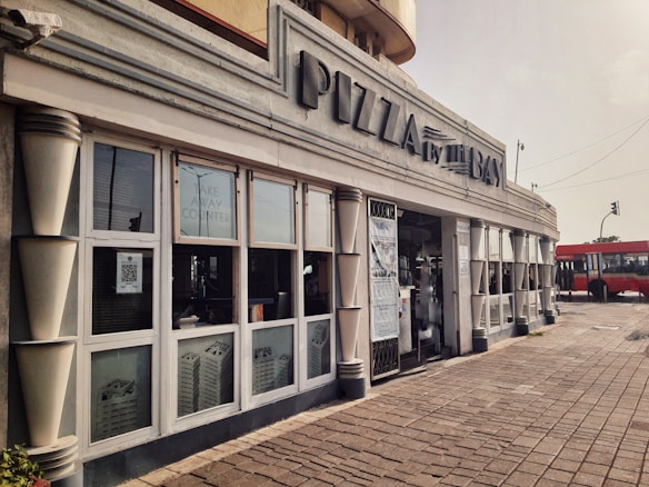 A restaurant with a streamlined facade featuring large windows and an art deco style. The signage reads 'PIZZA by the BAY,' and there is a take-away counter visible inside. The setting includes a tiled walkway and a red bus visible in the background, on a slightly overcast day.