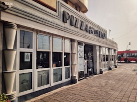 A restaurant with a streamlined facade featuring large windows and an art deco style. The signage reads 'PIZZA by the BAY,' and there is a take-away counter visible inside. The setting includes a tiled walkway and a red bus visible in the background, on a slightly overcast day.