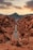A narrow road winds through a striking desert landscape with red rock formations on both sides. A vehicle travels along the stretch, surrounded by rugged terrain. In the background, a mountain range looms under a partly cloudy sky, illuminated by the warm glow of the sunset.