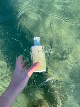 A hand is holding a bottle of volume shampoo partially submerged in clear, green-tinted water. The sunlight creates subtle reflections and ripples on the water surface.