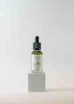 A small glass bottle with a dropper lid is positioned on a minimalist cube pedestal. The bottle contains a green liquid and has a label with floral designs and text, suggesting it is a natural hair oil product. The background is a soft, neutral color with a gentle gradient, enhancing the focus on the bottle.