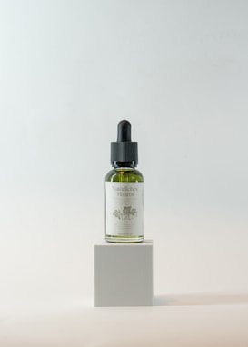 A small glass bottle with a dropper lid is positioned on a minimalist cube pedestal. The bottle contains a green liquid and has a label with floral designs and text, suggesting it is a natural hair oil product. The background is a soft, neutral color with a gentle gradient, enhancing the focus on the bottle.