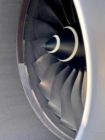 Close-up view of an aircraft jet engine's fan blades, showcasing the intricate and sleek design. The metallic blades are symmetrically arranged and reflect light, suggesting both power and precision.