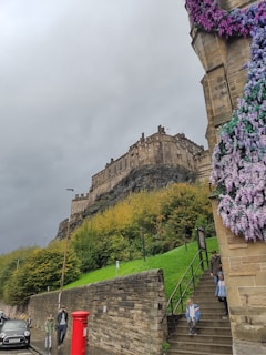 A historic castle sits atop a rocky hill, surrounded by lush green foliage and a cloudy sky. Below, two people walk along a street next to a red postbox, while a group of people, including children, descend a staircase. The corner of a building is adorned with cascading purple and white flowers.