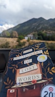 A book titled 'The Dictionary of Lost Words' is held up against a scenic background of a mountain landscape. The book has a dark cover with gold leaf patterns and a sticker indicating 'Reese's Book Club'. In the background, there is a clear view of green hills and a cloudy sky.
