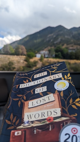 A book titled 'The Dictionary of Lost Words' is held up against a scenic background of a mountain landscape. The book has a dark cover with gold leaf patterns and a sticker indicating 'Reese's Book Club'. In the background, there is a clear view of green hills and a cloudy sky.