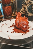 A realistic, sculpted heart made of a glossy, red material is placed on a white plate. Surrounding the heart are dramatic red splatters that resemble blood. In the background, various blurred statues and objects contribute to an artful, surreal display.