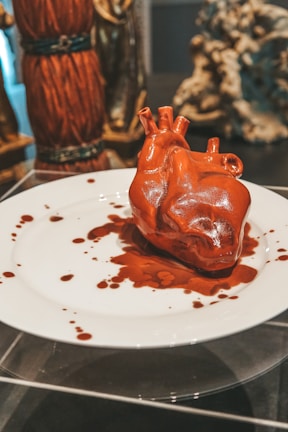 A realistic, sculpted heart made of a glossy, red material is placed on a white plate. Surrounding the heart are dramatic red splatters that resemble blood. In the background, various blurred statues and objects contribute to an artful, surreal display.