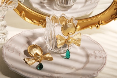 A set of ornate gold earrings with bow designs and green gemstone pendants are displayed on a delicate white plate with intricate patterns. In the background, a decorative golden mirror and crystal elements add an elegant touch, with soft, warm lighting creating a luxurious ambience.
