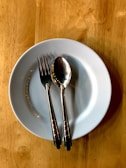 A white ceramic plate sits on a wooden table with a stainless steel fork and spoon placed on it, arranged parallel to each other.