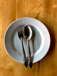 A white ceramic plate sits on a wooden table with a stainless steel fork and spoon placed on it, arranged parallel to each other.