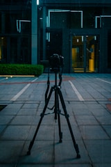 A camera is mounted on a tripod positioned on a tiled pavement in an urban area. Behind it, there is a modern building with reflective glass and bright blue neon lights outlining parts of the architecture. A small green hedge is visible to the left.