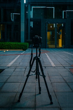 A camera is mounted on a tripod positioned on a tiled pavement in an urban area. Behind it, there is a modern building with reflective glass and bright blue neon lights outlining parts of the architecture. A small green hedge is visible to the left.