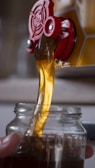 Golden liquid, likely honey, pouring from a red spout into a clear glass jar. The honey flows smoothly, catching the light and creating a rich, warm color.