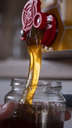 Golden liquid, likely honey, pouring from a red spout into a clear glass jar. The honey flows smoothly, catching the light and creating a rich, warm color.