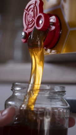 Golden liquid, likely honey, pouring from a red spout into a clear glass jar. The honey flows smoothly, catching the light and creating a rich, warm color.