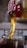 Golden liquid, likely honey, pouring from a red spout into a clear glass jar. The honey flows smoothly, catching the light and creating a rich, warm color.