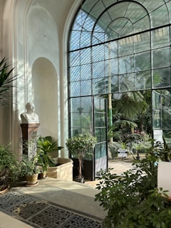 A bright conservatory with large arched windows allowing sunlight to stream in, illuminating various green plants and trees. A marble bust on a pedestal stands among the foliage, creating a classical atmosphere. The room is filled with lush greenery, and the patterned metal floor grating adds a decorative element.