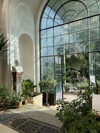 A bright conservatory with large arched windows allowing sunlight to stream in, illuminating various green plants and trees. A marble bust on a pedestal stands among the foliage, creating a classical atmosphere. The room is filled with lush greenery, and the patterned metal floor grating adds a decorative element.