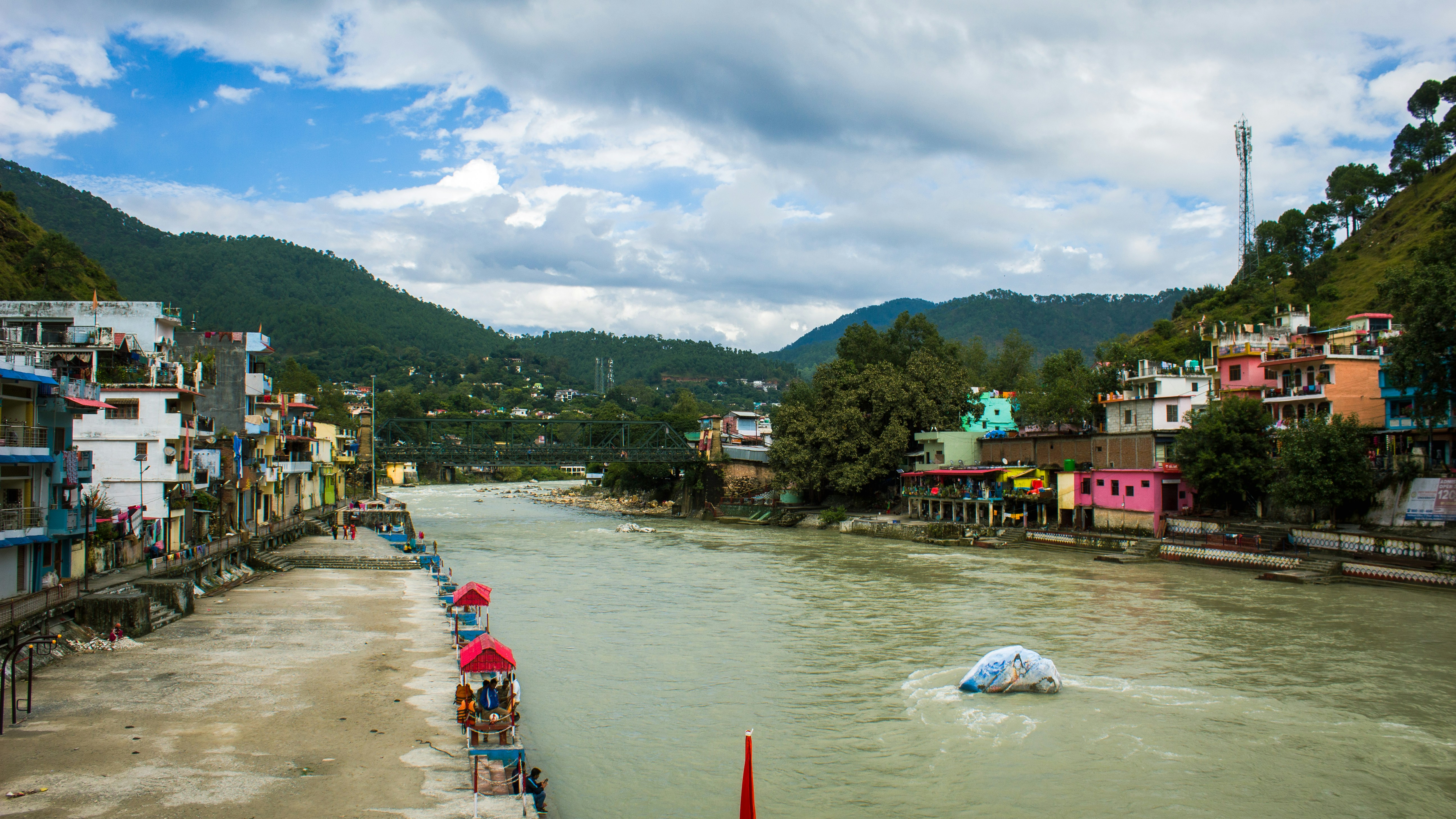 A wide shot of the Ganga river flowing through a bustling city in Uttarakhand, with some visible signs of human activity along the banks and a hint of a discharge pipe in the distance.