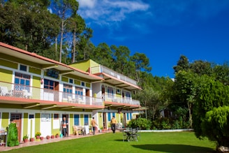 A two-story building with green and white exterior nestled among tall trees, with a well-maintained lawn in the foreground. Several potted plants line the exterior, and a few people are present, some standing and others walking. The sky is clear and vivid blue, providing a bright and cheerful backdrop.