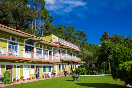 A two-story building with green and white exterior nestled among tall trees, with a well-maintained lawn in the foreground. Several potted plants line the exterior, and a few people are present, some standing and others walking. The sky is clear and vivid blue, providing a bright and cheerful backdrop.