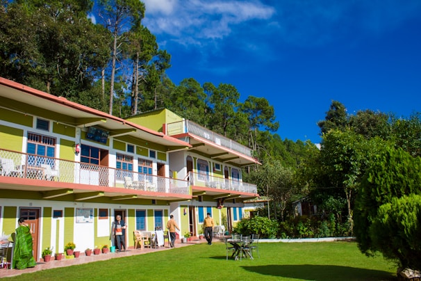 A two-story building with green and white exterior nestled among tall trees, with a well-maintained lawn in the foreground. Several potted plants line the exterior, and a few people are present, some standing and others walking. The sky is clear and vivid blue, providing a bright and cheerful backdrop.
