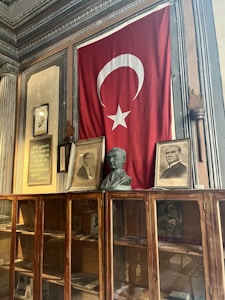 A large Turkish flag with a white star and crescent hangs on the wall. Below it, there are framed photographs and a bust on top of a glass-fronted wooden cabinet. The walls have ornate details with decorative moldings.