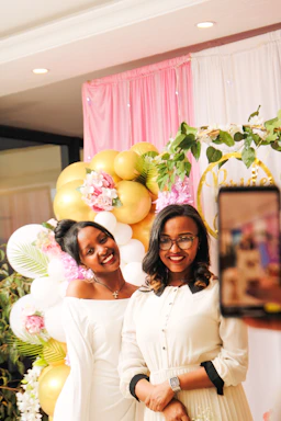 A joyful quinceañera girl posing with friends in front of a pink and gold photo booth backdrop.