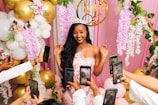 A smiling woman wearing a tiara and a sash that reads 'Bride to Be' stands surrounded by pink and white floral decorations and golden balloons. Multiple hands holding smartphones are pointed towards her, capturing photos.