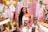 A smiling woman wearing a tiara and a sash that reads 'Bride to Be' stands surrounded by pink and white floral decorations and golden balloons. Multiple hands holding smartphones are pointed towards her, capturing photos.