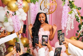 A smiling woman wearing a tiara and a sash that reads 'Bride to Be' stands surrounded by pink and white floral decorations and golden balloons. Multiple hands holding smartphones are pointed towards her, capturing photos.